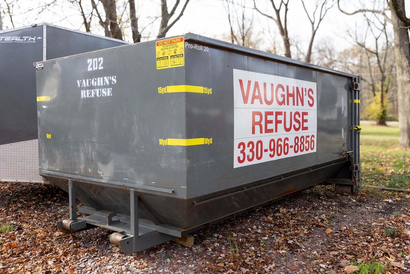 Large Vaughn's Refuse gray dumpster with company logo, phone number, and caution signs, located in an outdoor wooded area with fallen leaves.