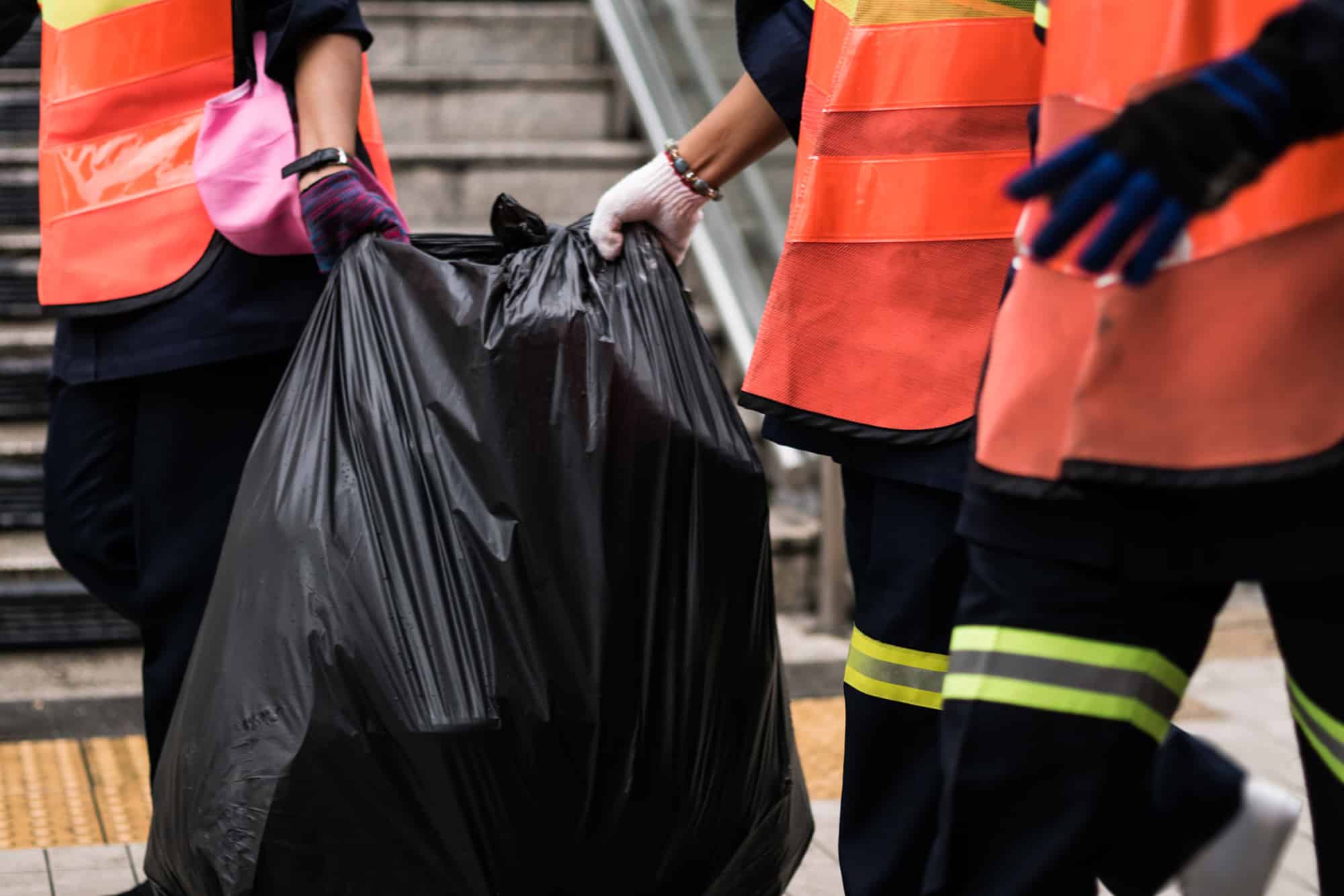 Workers wearing reflective orange vests and gloves carrying a large black garbage bag, demonstrating efficient junk removal services provided by Vaughn's Refuse.