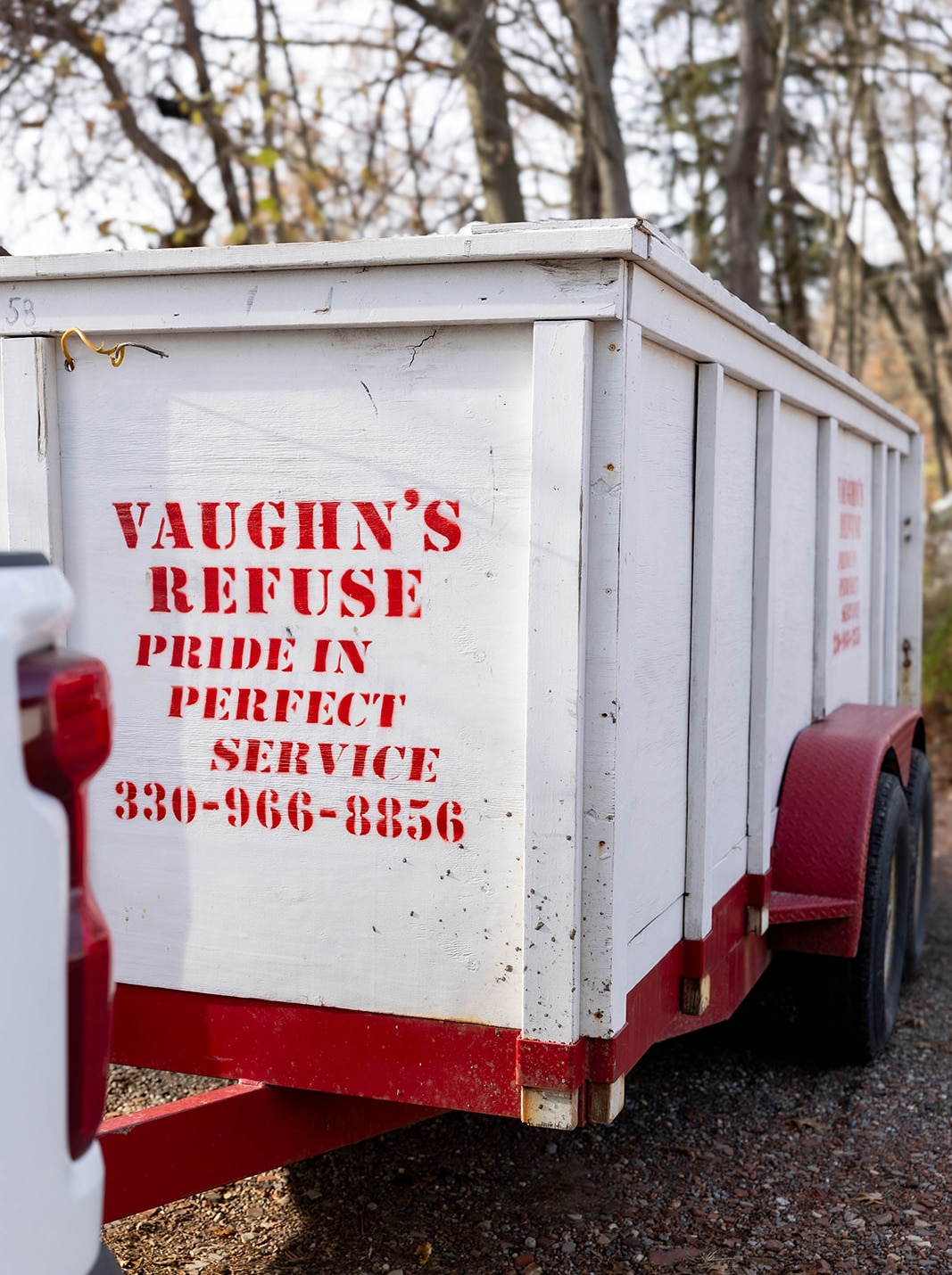 Close-up of Vaughn's Refuse white and red trailer featuring company logo, slogan, and contact number, parked on a gravel surface with trees in the background.