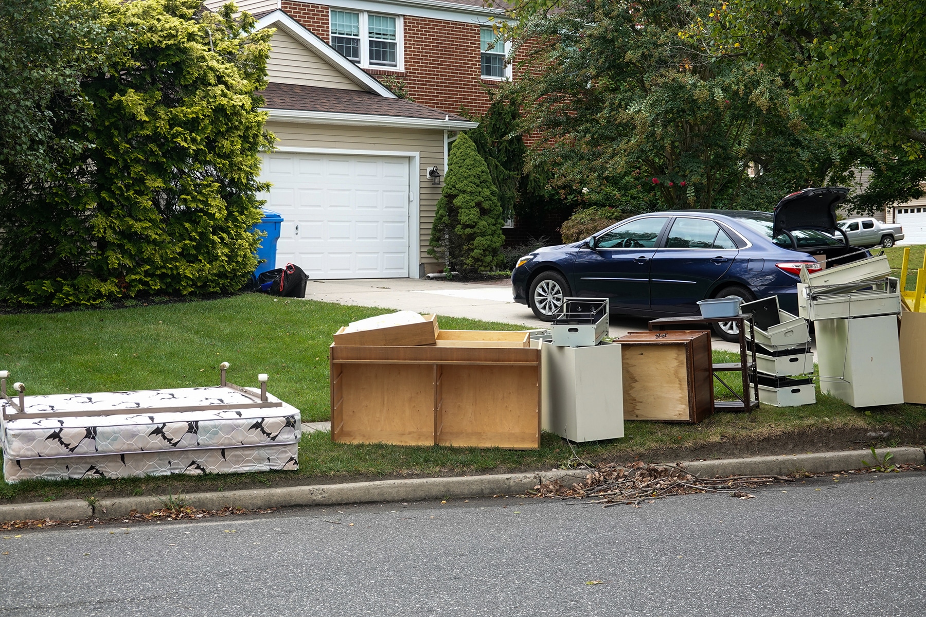  Junk removal in progress outside a suburban home, with an old mattress, broken dressers, filing cabinets, and various discarded furniture pieces lined up along the curb for pickup. A blue car with an open trunk is parked in the driveway, suggesting a major cleanout.