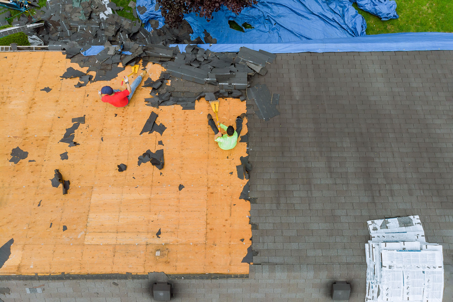 Aerial view of two workers removing damaged roof shingles from a house during roof repair, with tarps and debris scattered nearby.